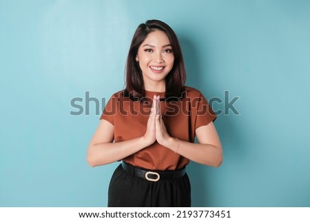 Smiling young Asian woman gesturing traditional greeting isolated over blue background