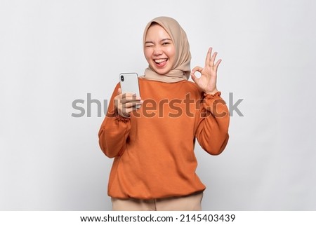 Smiling young Asian woman in orange shirt holding mobile phone and showing okay sign isolated over white background