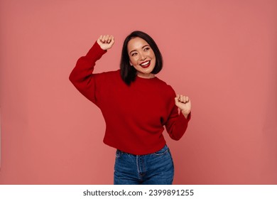 Smiling young asian woman in red sweater dancing isolated over pink studio background - Powered by Shutterstock