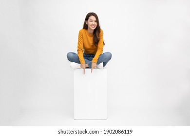 Smiling Young Asian Woman Sitting On White Box And Pointing Down Empty Copy Space Isolated On White Background