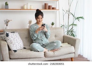 smiling young asian woman sitting on comfortable coach in living room, using mobile phone applications, shopping, playing online games, chatting with friends, watching funny videos with pajama