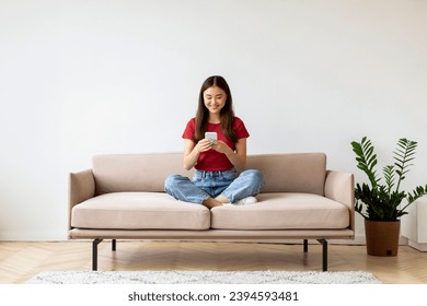 Smiling Young Asian Woman With Smartphone Relaxing On Couch At Home, Beautiful Korean Female Resting On Sofa And Using Mobile Phone, Texting With Friends Or Shopping Online, Copy Space - Powered by Shutterstock