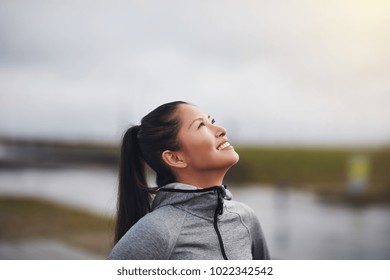 Smiling Young Asian Woman In Sportswear Standing Outside Looking Up At The Sky While Out For A Jog