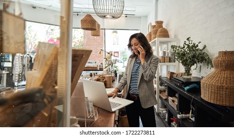 Eine junge Asiatin, die hinter einer Theke steht, in ihrer stilvollen Boutique, die an einem Laptop arbeitet und auf einem Handy spricht