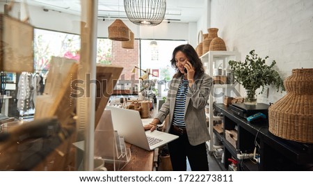 Smiling young Asian woman standing behind a counter in her stylish boutique working on a laptop and talking on a cellphone