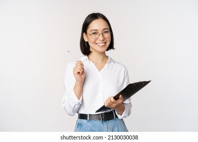 Smiling young asian woman taking notes with pen on clipboard, looking happy, standing against white background