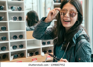 Smiling Young Asian Woman Traveler Trying On Sunglasses On Mirror In Optician Face Looking Camera. Female Buying Glasses Shop Store On Bargain Summer Sale On Hollywood Boulevard Street Mall Centre