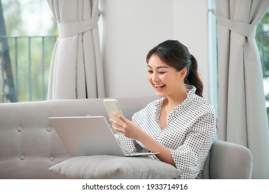 Smiling Young Asian Woman Using Mobile Phone While Sitting On A Couch At Home With Laptop Computer