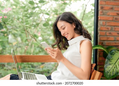 Smiling Young Asian Woman Using Mobile Phone While Sitting On A Couch At Home With Laptop Computer