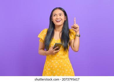 Smiling young Asian woman using a mobile phone and pointing fingers up with an idea on purple background