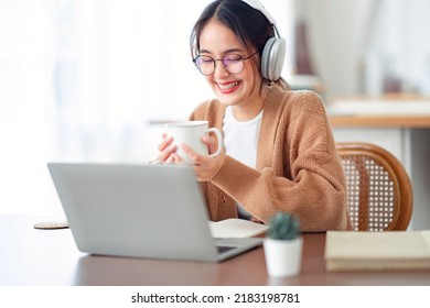 Smiling young asian woman using laptop web camera while holding a cup of coffee and wearing headphones in the kitchen at home.