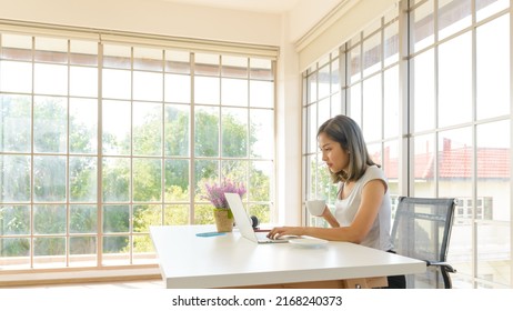 Smiling Young Asian Woman Wear Casual Clothing Working With Computer Laptop And Holding Coffee Mug In Living Room At Home. WFH. Work At Home Concept