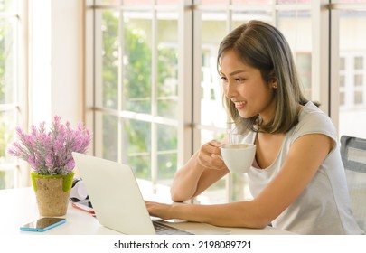 Smiling Young Asian Woman Wear Casual Clothing Working With Computer Laptop And Holding Coffee Mug In Living Room At Home. WFH. Work At Home Concept