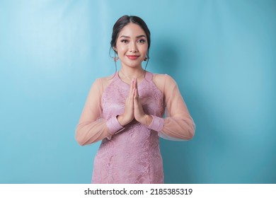 Smiling young Asian woman wearing modern kebaya gesturing greeting or namaste isolated over blue background