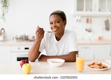 Smiling Young Attractive Black Woman Eating Porridge Sits At Table With Cookies, Juice And Apples In Scandinavian Kitchen Interior. Good Morning, Health Care, Proper Nutrition And Breakfast At Home