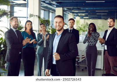 Smiling young attractive boss and business leader man proudly looking at camera with team of people applauding in background in office. Business team or group of staff congratulating their leader. - Powered by Shutterstock