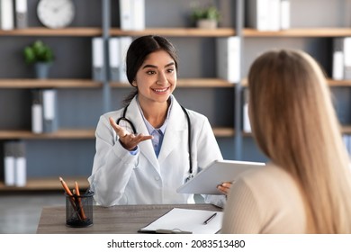 Smiling young attractive indian lady medical worker in white coat gestures and consultation patient at workplace in clinic office interior. Modern medicine, treatment and therapy with family doctor