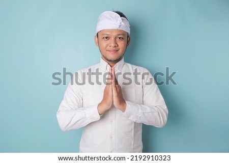 Smiling young Balinese man wearing udeng or traditional headband and white shirt gesturing greeting or namaste isolated over blue background