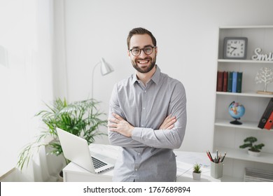 Smiling Young Bearded Business Man In Gray Shirt Glasses Standing Near Desk With Laptop Computer In Light Office On White Wall Background. Achievement Business Career Concept. Holding Hands Crossed