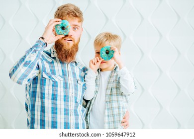 Smiling young bearded hipster man with his cute son having fun while holding donuts on their eyes over white polygonal background.