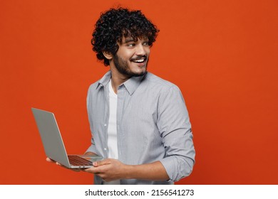 Smiling Young Bearded Indian Man 20s Years Old Wears Blue Shirt Hold Use Work On Laptop Pc Computer Typing Browsing Chatting Send Sms Looking Behind Isolated On Plain Orange Background Studio Portrait