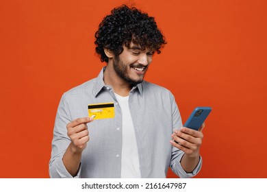 Smiling Young Bearded Indian Man 20s Years Old Wears Blue Shirt Using Mobile Cell Phone Hold Credit Bank Card Doing Online Shopping Order Delivery Isolated On Plain Orange Background Studio Portrait