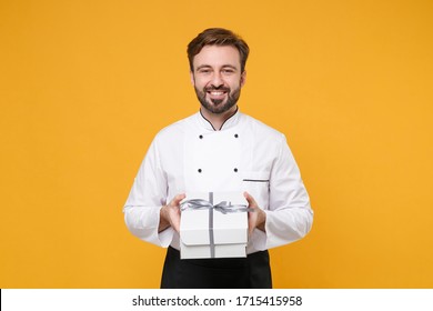 Smiling Young Bearded Male Chef Cook Or Baker Man In White Uniform Shirt Posing Isolated On Yellow Wall Background. Cooking Food Concept. Mock Up Copy Space. Hold Present Box With Gift Ribbon Bow