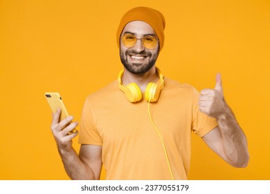 Smiling young bearded man 20s in basic casual t-shirt headphones eyeglasses hat standing using mobile cell phone typing sms message showing thumb up isolated on yellow background, studio portrait - Powered by Shutterstock