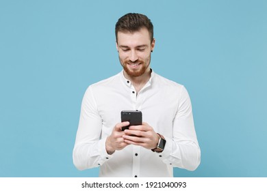 Smiling young bearded man guy 20s in white classic shirt isolated on pastel blue wall background studio portrait. People lifestyle concept. Mock up copy space. Using mobile phone, typing sms message
