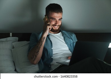 Smiling Young Bearded Man Laying On Bed At Home At Night, Talking On Mobile Phone While Working On Laptop Computer