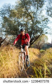Smiling Young Bearded Man In Red Long Sleeve Cycling Jersey Riding Mountain Bike Along A Path Through Tall Grass. Low Angle Front View.