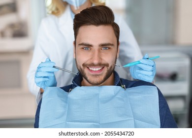 Smiling young bearded man sitting in dental chair, visiting modern dental clinic, having regular checkup, unrecognizable woman dentist holding dental tools next to male patient mouth - Shutterstock ID 2055434720
