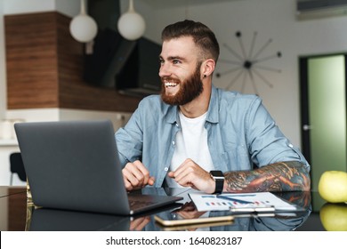 Smiling young bearded man working on laptop computer while sitting at the kitchen table