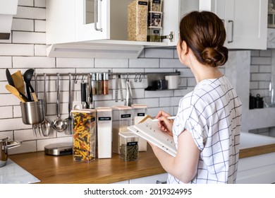 Smiling young beautiful business female taking notes in paper diary at modern Nordic style kitchen. Happy woman writing in notepad surrounded by comfortable organization storage minimalistic cuisine - Powered by Shutterstock