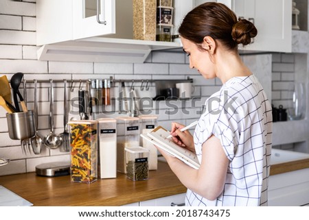 Smiling young beautiful business female taking notes in paper diary at modern Nordic style kitchen. Happy woman writing in notepad surrounded by comfortable organization storage minimalistic cuisine