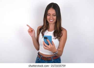 Smiling young beautiful caucasian woman wearing white top over white background pointing finger at blank space holding phone in one hand