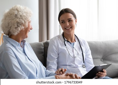 Smiling Young Beautiful Female Doctor Nurse In Medical Uniform Enjoying Pleasant Conversation With Happy Older Hoary Woman, Consulting Mature Patient At Home Visit, Sitting Together On Sofa.