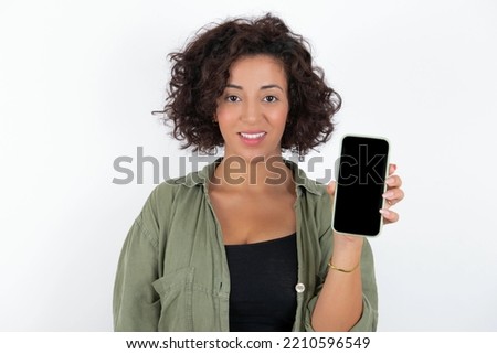 Smiling young beautiful woman with curly short hair wearing green overshirt over white wall Mock up copy space. Hold mobile phone with blank empty screen