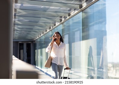 Smiling Young Biracial Businesswoman Talking On Phone Walking With Luggage At Airport Corridor. Unaltered, Technology, Occupation, Business Travel And Transportation Concept.