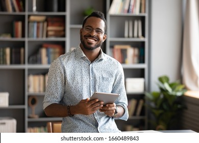 Smiling young biracial man in glasses holding digital computer tablet, lookig at camera. Addicted to technology, happy handsome businessman using electronic device, web surfing internet indoors.