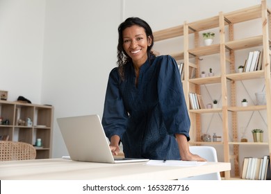 Smiling young biracial woman standing at table with computer, looking at camera. Happy african american business woman company employee manager professional posing for photo alone in modern office.