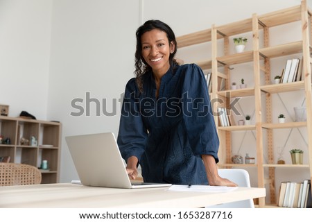 Smiling young biracial woman standing at table with computer, looking at camera. Happy african american business woman company employee manager professional posing for photo alone in modern office.