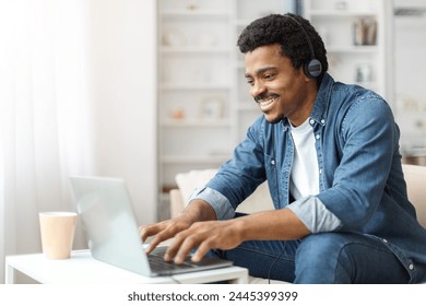 Smiling young black adult male in a denim jacket using laptop at a white desk at home with a coffee cup beside him. - Powered by Shutterstock