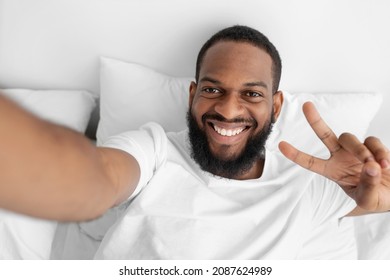 Smiling young black bearded man lies on white bed in bedroom, takes selfie and shows peace sign with his hand. Covid-19 lockdown, social distancing, rest and relax at home, livestream and video call