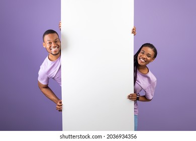 Smiling young black couple holding white vertical advertisement board, demonstrating free copy space for your text or design, positive guy and lady peeking out banner, purple violet background - Shutterstock ID 2036934356