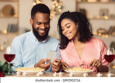 Smiling Young Black Couple Looking At Smartphone And Having Dinner. African American Woman And Man Sharing Social Media On Smart Phone, Eating Pasta During Romantic Date, Sitting At Table