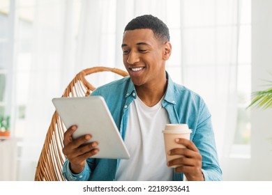 Smiling Young Black Guy In Casual Chatting On Tablet, Watch Video, Enjoy Takeaway Drink, Sitting In Armchair In Bright Room Interior. Rest At Spare Time, Coffee Break With New App, Surfing In Internet