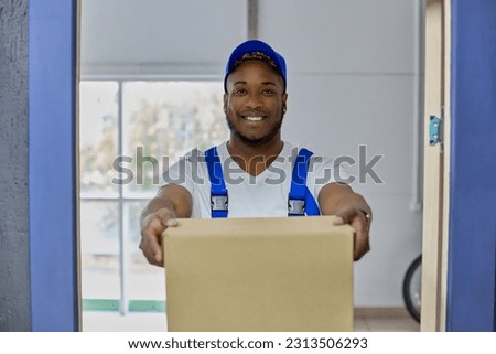 Smiling young black guy delivers parcel in blue uniform in cardboard box to customer's house. Professional delivery service employee of a man in a blue cap qualitatively and quickly performs his work Stock foto © 