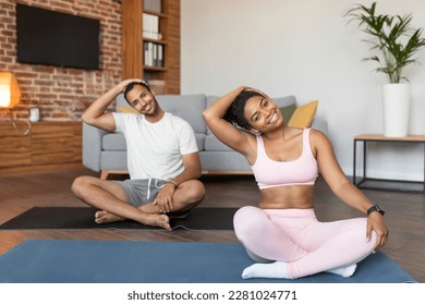 Smiling young black guy and lady in sportswear doing exercises, practicing yoga, stretching muscles in lotus position on mat together in living room interior. Workout, health care and fitness at home - Powered by Shutterstock