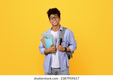 Smiling young black male student in glasses carrying notebooks and backpack stands against yellow background and smiling at camera - Powered by Shutterstock